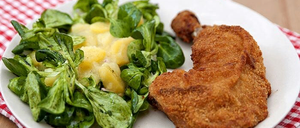 Ein weißer Teller mit einem rot-weiß karierten Tuch enthält einen grünen Spinatsalat mit Ananas und einer gebratenen Hühnerschnitzel.