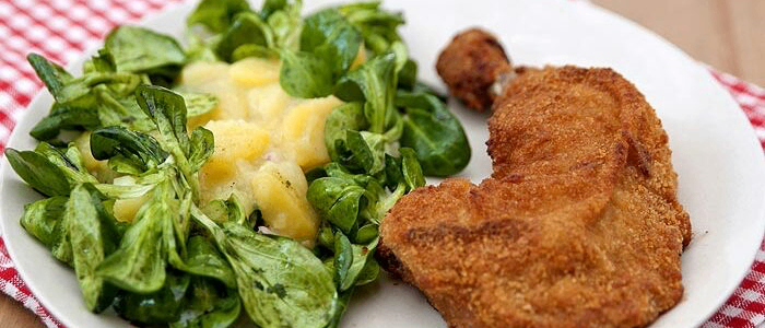 Ein weißer Teller mit einem rot-weiß karierten Tuch enthält einen grünen Spinatsalat mit Ananas und einer gebratenen Hühnerschnitzel.