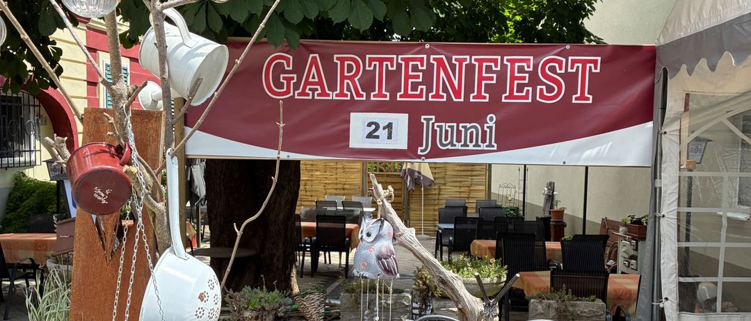 Ein Gartenbereich mit Topfpflanzen, einem Banner mit der Aufschrift 'Gartenfest 21 Juni' und einem Baum mit hängenden Dekorationsartikeln.