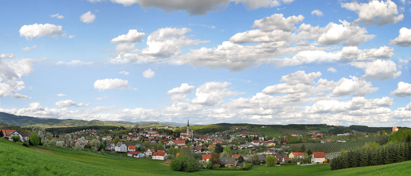 Ein kleines Dorf mit roten Dächern und einem Kirchturm in der Ferne, vor einer Kulisse aus grünen Hügeln und einem blauen Himmel mit verstreuten Wolken.