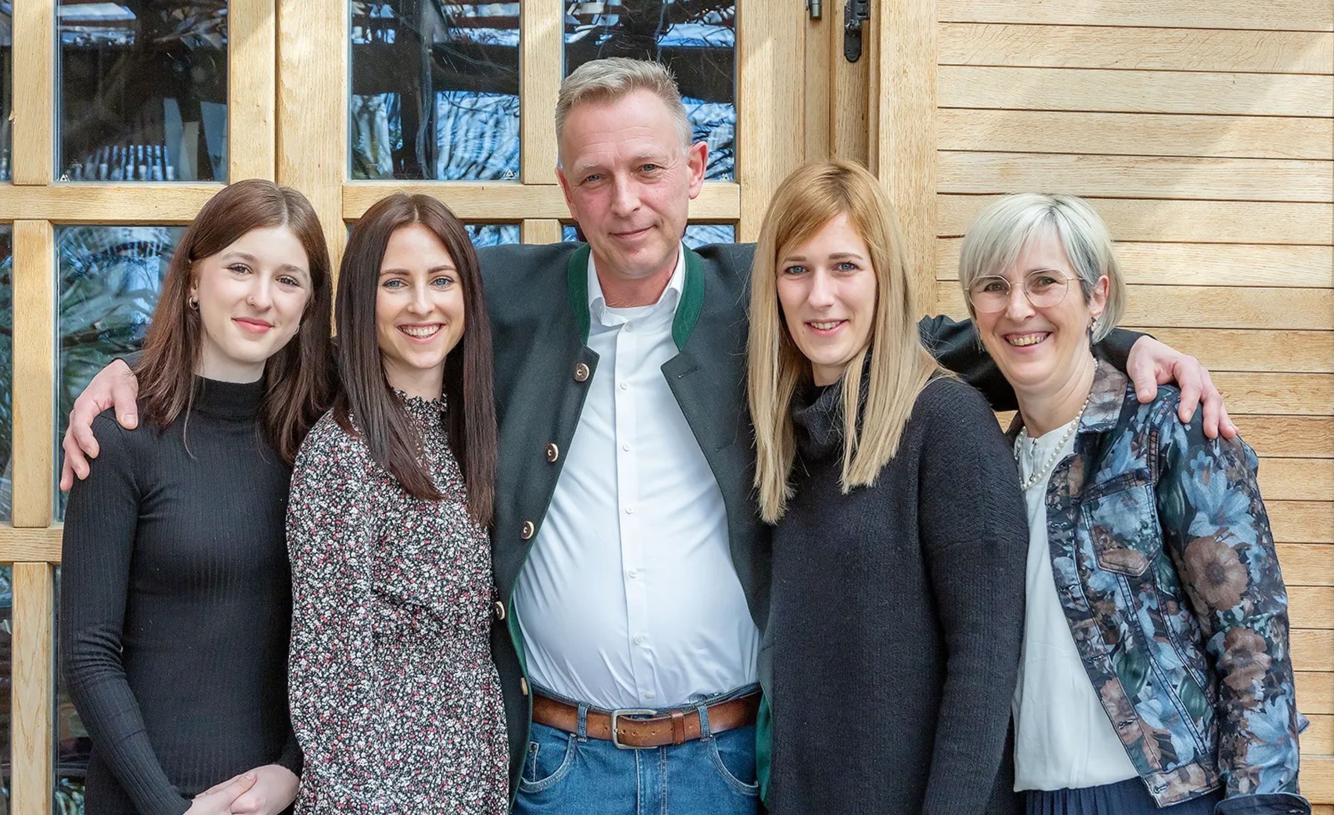 Eine fünfköpfige Familie steht lächelnd vor einer Holztür. Der Mann hat seinen Arm um die Schultern zweier Frauen gelegt. Dahinter reflektiert ein Glasfenster Bäume.