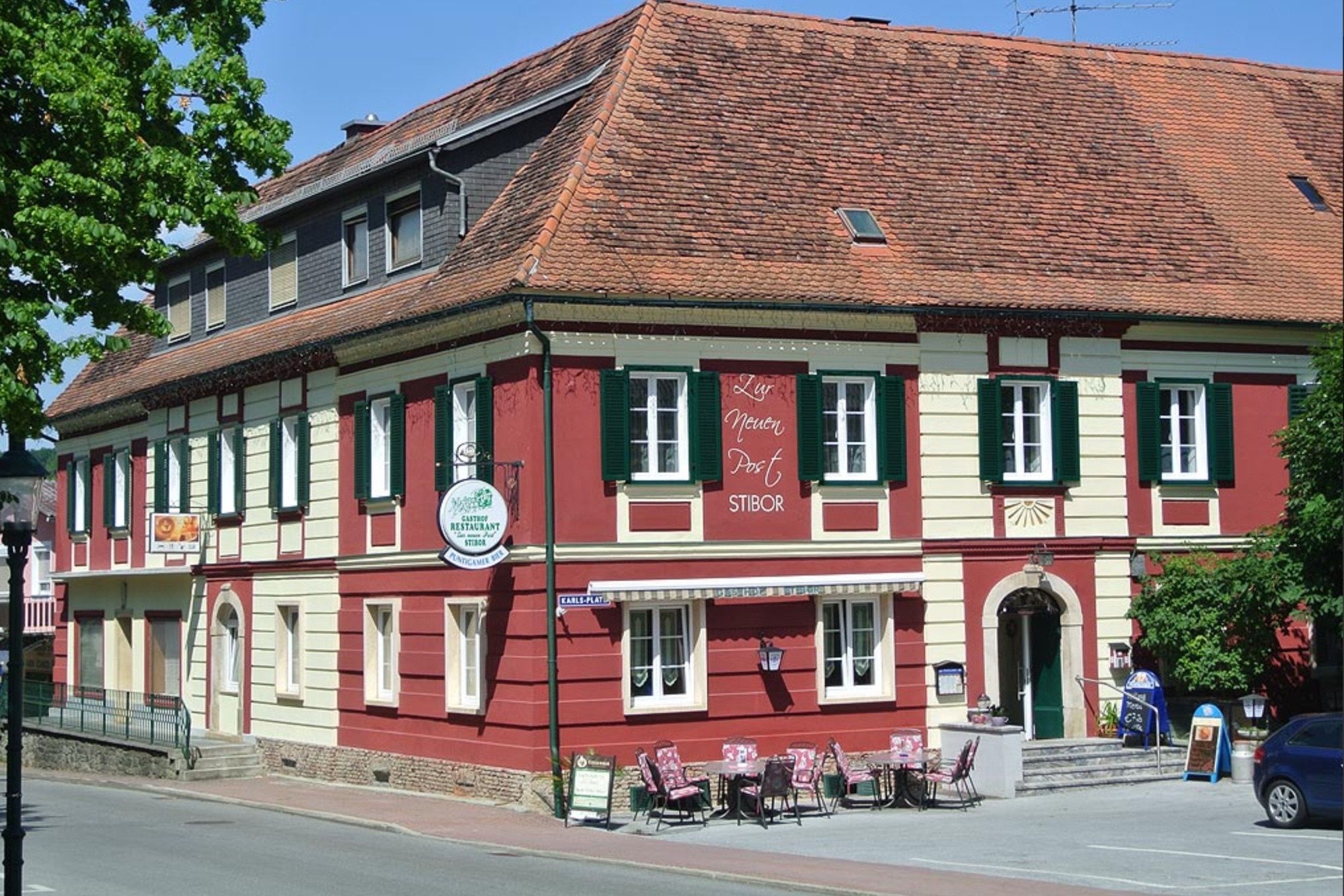 Ein rotes Gebäude mit weißen Akzenten und grünen Fensterläden trägt ein Schild mit der Aufschrift 'Lys neuer Post STIBOR'. Draußen sind Tische und Stühle für Kunden aufgestellt, und ein rundes Schild mit 'Café Restaurant STIBOR' hängt an der Wand.