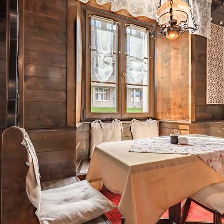 Ein hölzerner Raum mit einem mit einem Blumentischtuch bedeckten Esstisch. Es gibt eine von der Decke hängende Lampe und ein Fenster mit Vorhängen.