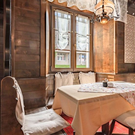 Ein hölzerner Raum mit einem mit einem Blumentischtuch bedeckten Esstisch. Es gibt eine von der Decke hängende Lampe und ein Fenster mit Vorhängen.