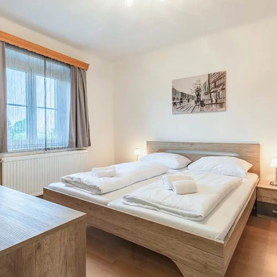 Ein Schlafzimmer mit einem Holzbett, zwei Kissen und einer Lampe auf dem Nachttisch. Der Raum hat weiße Wände und ein Fenster mit Vorhängen.