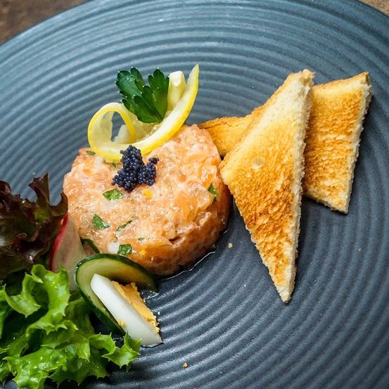 Ein Teller mit Lachstatar mit Kaviar, Zitrone und Toast, begleitet von grünem Salat und Gurkenscheiben auf einem blauen Teller.
