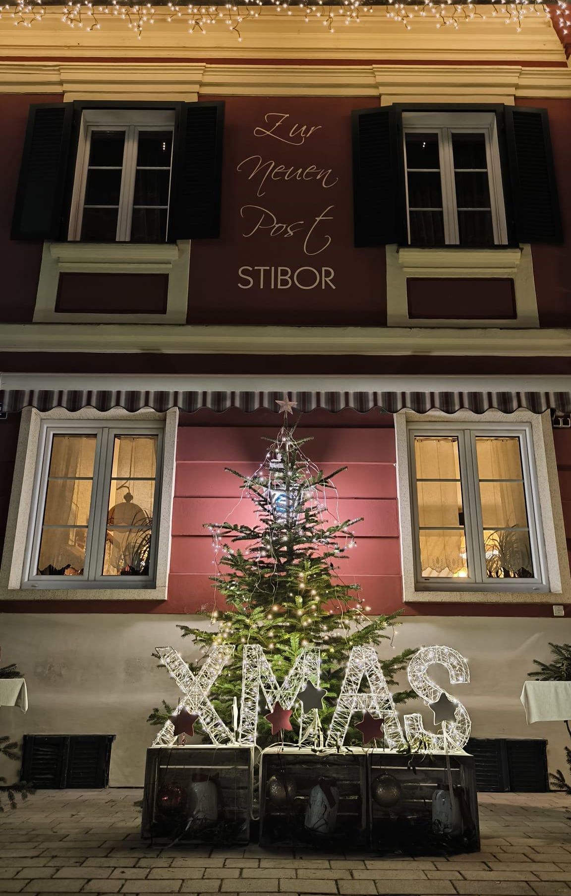 Ein Weihnachtsbaum steht vor einem Gebäude mit dem Schild 'Newen Post' darüber. Der Baum ist mit Lichtern und einem Stern geschmückt. Es gibt zwei Fenster auf beiden Seiten des Baumes.