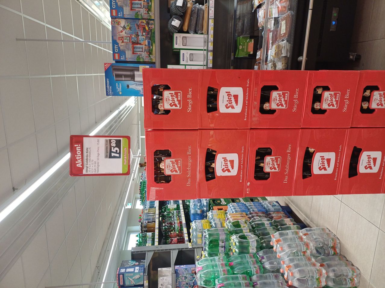Ein Supermarktregal mit roten Stiegl-Bierkästen auf einem Regal gestapelt. Jeder Kasten enthält sechs Flaschen. Ein Preisschild zeigt einen Rabatt, wobei der ursprüngliche Preis 15,80 Euro betrug.