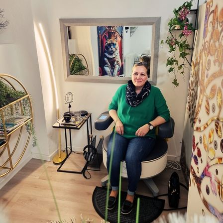 Eine Frau sitzt in einem Stuhl in einem Raum mit einem Spiegel und einer Blumenwanddekoration. Sie trägt ein grünes Shirt, Jeans und einen Schal.