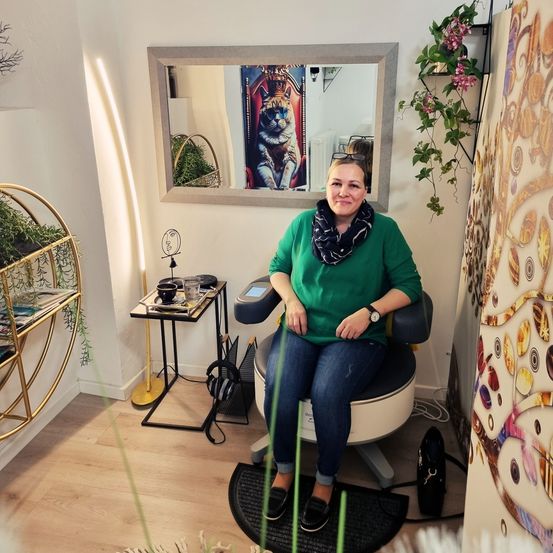 Eine Frau sitzt in einem Stuhl in einem Raum mit einem Spiegel und einer Blumenwanddekoration. Sie trägt ein grünes Shirt, Jeans und einen Schal.