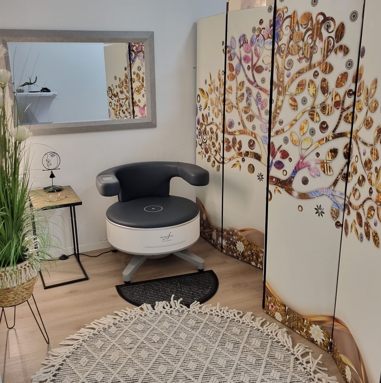 A small room with a white chair and a large mirror. A plant in a basket is on the left. A carpet and a tree-printed screen are on the right.