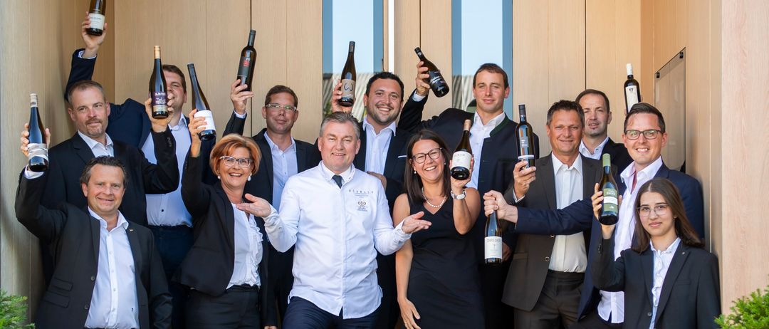 A group of people, dressed in suits and dresses, are posing for a photo on the stairs. They are holding wine bottles. The wooden sign with the word DiVA is placed on the stairs.