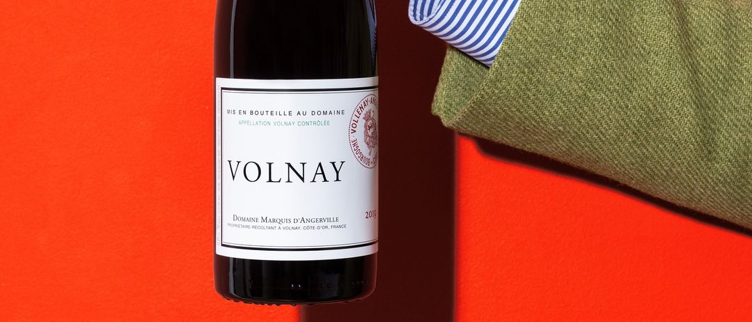 Eine Person hält eine Flasche Volnay-Wein vor einem leuchtend roten Hintergrund. Das Etikett zeigt Volnay, Domaine Marquis d'Angerville.