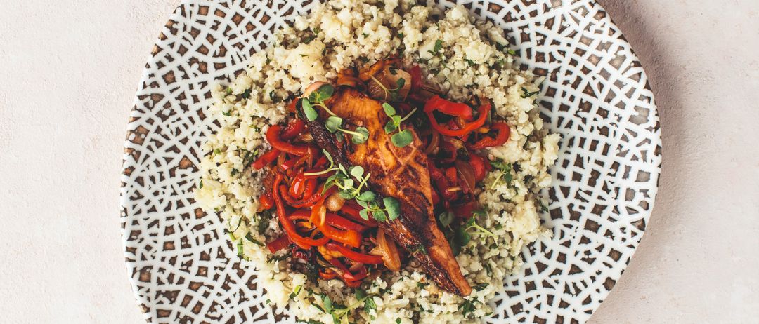 Ein Teller mit geometrischem Design hält ein Gericht aus Couscous mit roten Paprikaschoten und Fleisch, wahrscheinlich einem Hähnchenschenkel.