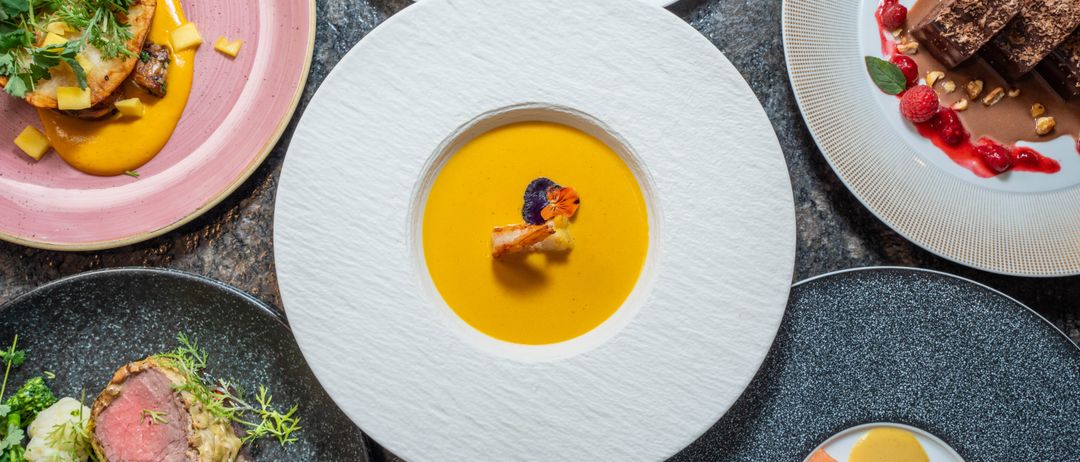 Ein Tisch zeigt verschiedene Gourmet-Gerichte. Eine zentrale Platte zeigt eine cremige Suppe mit einer dekorativen Garnierung. Umgebende Teller zeigen verschiedene farbenfrohe Gerichte mit Garnierungen.
