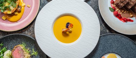 Ein Tisch zeigt verschiedene Gourmet-Gerichte. Eine zentrale Platte zeigt eine cremige Suppe mit einer dekorativen Garnierung. Umgebende Teller zeigen verschiedene farbenfrohe Gerichte mit Garnierungen.