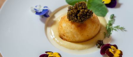 Ein weißer Teller hält eine goldbraune kugelförmige Speise, die mit schwarzem Kaviar bedeckt ist, umgeben von cremiger Sauce und mit grünen Blättern und bunten Blumen garniert.