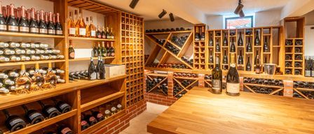 Ein Weinkeller mit Holzregalen und Ziegelwänden. Die Regale sind mit Weinflaschen gefüllt, darunter auch Champagner. Auf dem Tisch in der Mitte stehen Weinflaschen. Es gibt Strahler an der Decke.