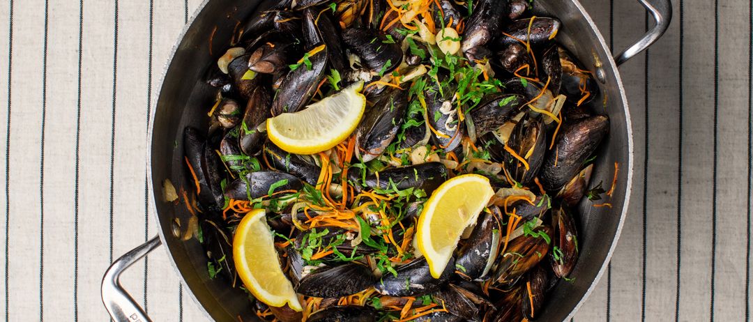 Eine Pfanne mit Muscheln, garniert mit Zitronenscheiben und Kräutern, auf einer gestreiften Oberfläche mit einem Teller Brot in der Nähe.