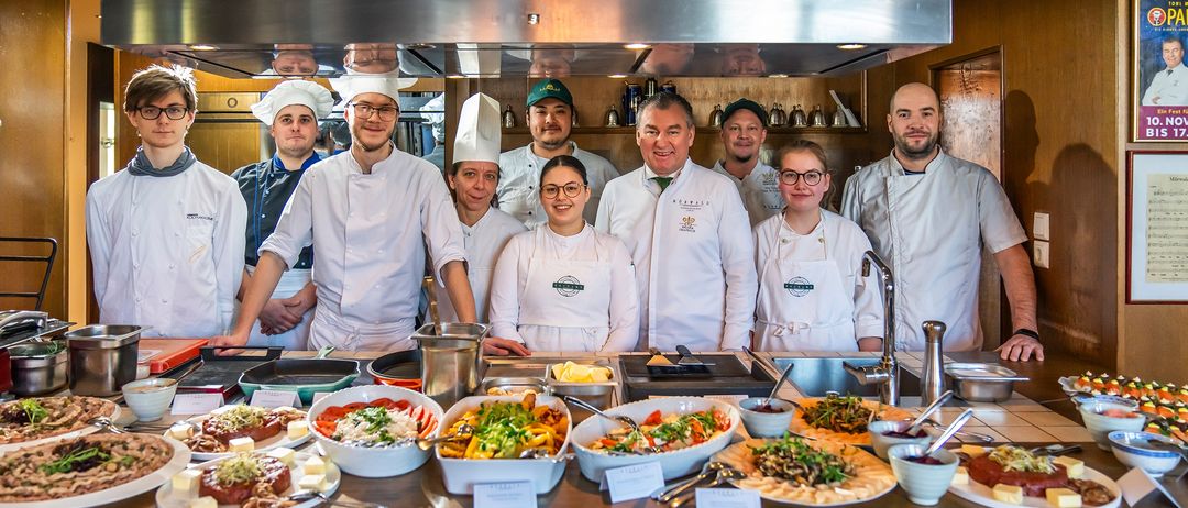 Eine Gruppe von Köchen in einer Küche, lächelnd und für ein Foto posierend. Eine Vielzahl von Gerichten steht auf dem Tresen, darunter Salate, Sandwiches und andere Speisen.