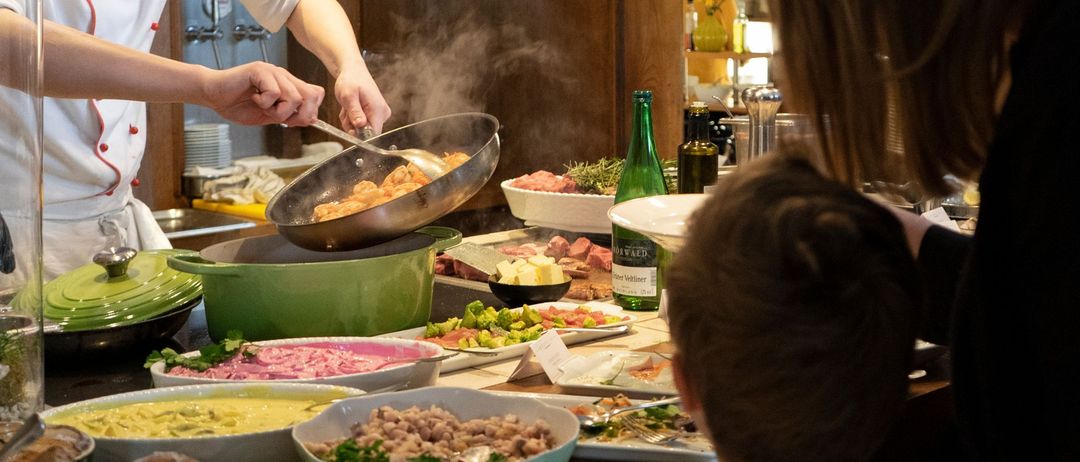 Ein Koch bereitet ein Buffet zu, an dem eine Familie sitzt. Verschiedene Gerichte und Flaschen stehen auf dem Tisch, und eine Frau steht hinter der sitzenden Familie.