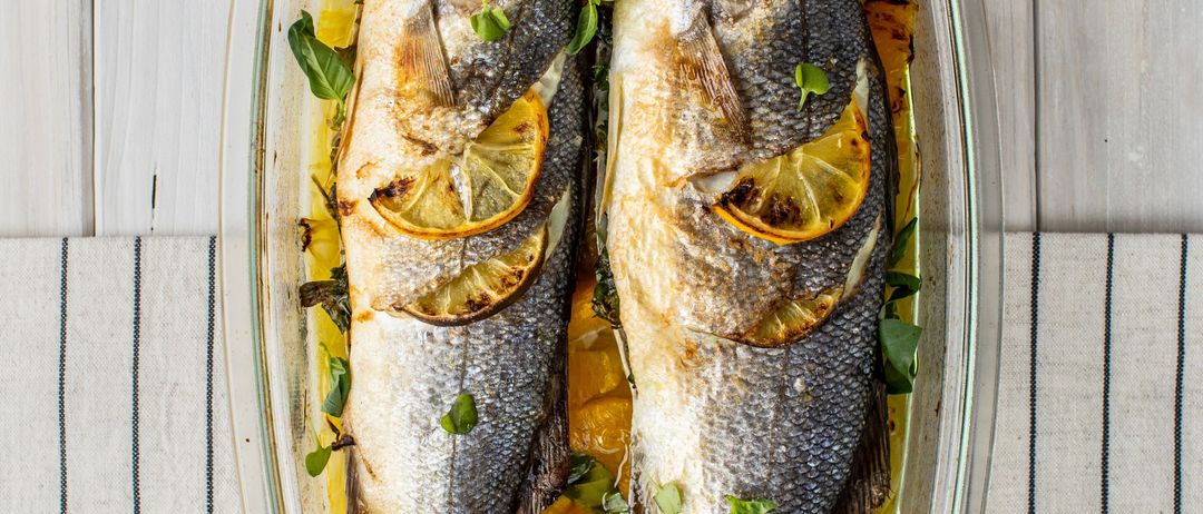 Zwei gegrillte Fische mit Zitronen und Kräutern in einer Glasschale auf einer weißen Oberfläche.