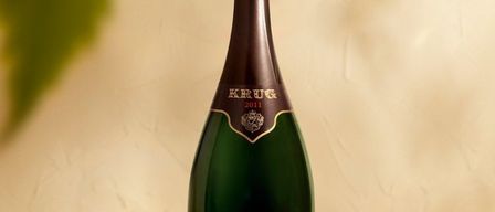 Eine Flasche Krug Champagner von 2011 wird auf einer weißen Oberfläche mit einem grünen Blatt im Hintergrund ausgestellt.