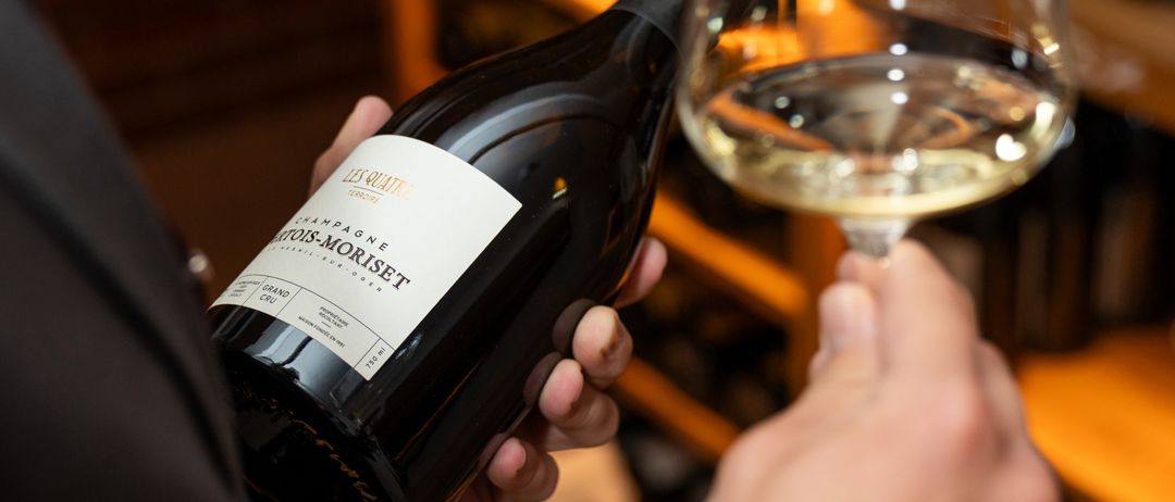 Eine Person in einem schwarzen Anzug hält eine Flasche Champagner, mit einem Glas gefüllt mit Weißwein.