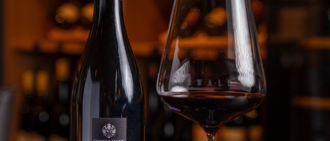 Eine Flasche Rotwein mit dem Etikett '2021 hochberg Blaufränkisch' steht neben einem gefüllten Weinglas. Dahinter befindet sich ein Holzregal mit weiteren Weinflaschen.