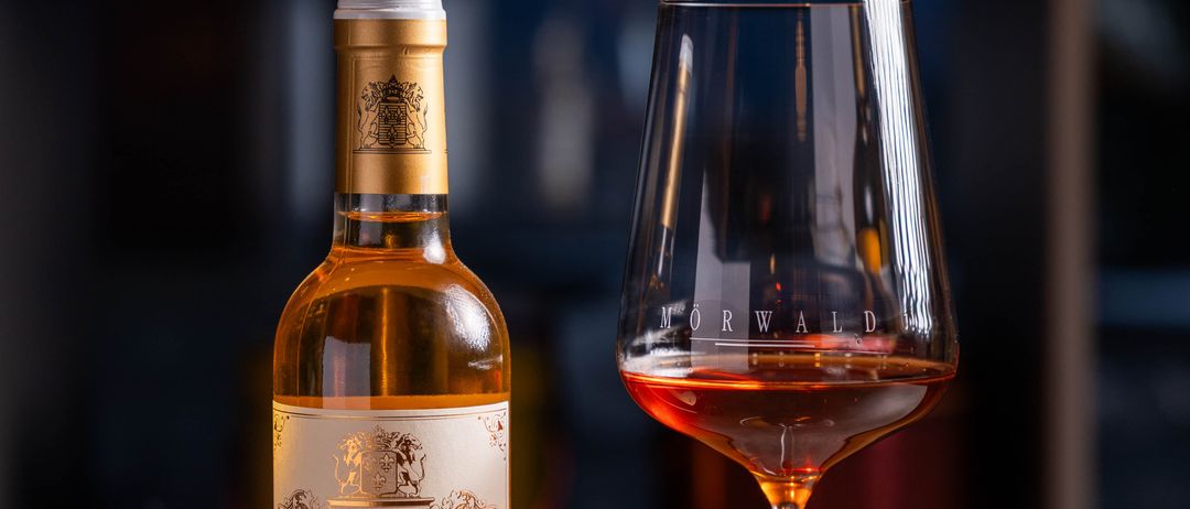 Eine Flasche Weißwein von Chateau Coutet steht neben einem Weinglas mit dem Label 'Morwald'. Dahinter befindet sich ein Regal mit verschiedenen Flaschen.