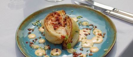A plate of roasted cabbage with bacon and sauce sits on a white table, accompanied by two glasses of water and a wine glass.