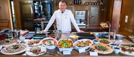 A chef in a white jacket stands behind a table with various dishes, smiling for the camera. The table is filled with multiple plates of food, including salads, meats, and vegetables. Behind him, a kitchen with stainless steel appliances and wooden shelves is visible.