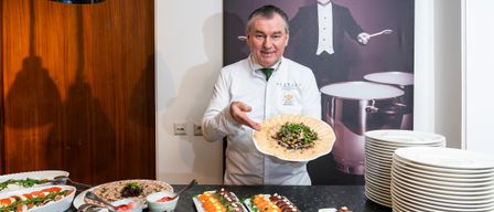 A chef stands before a table with various dishes, including sushi and other foods. In the background, a large portrait of a man in formal attire is visible.