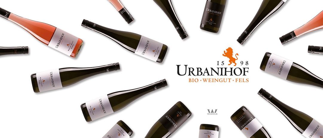 Eine Reihe von Weinflaschen in einem Kreis angeordnet, mit dem Urbanihof-Logo und Markennamen deutlich sichtbar. Die Flaschen sind in verschiedenen Größen und Formen, und das Bild zeigt ein Löwenemblem.