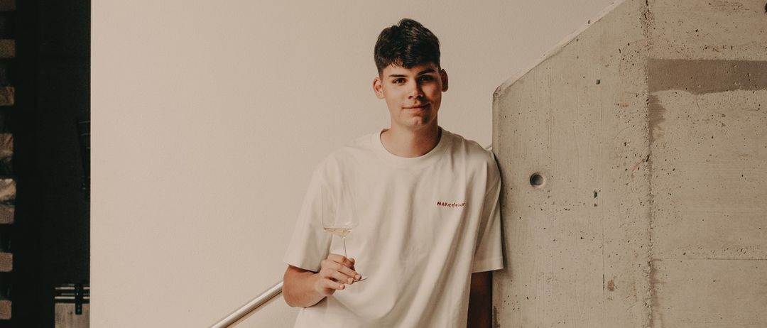 Ein junger Mann steht auf der Treppe, hält ein Weinglas in der rechten Hand und lehnt sich gegen eine Betonwand. Er trägt ein weißes T-Shirt und Shorts.