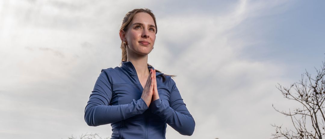 Eine Frau in blauer und brauner Yogakleidung führt eine Yoga-Pose auf einer Holzterrasse aus. Hinter ihr steht ein roter Sofa, ein Glaszaun und ein blattloser Baum.