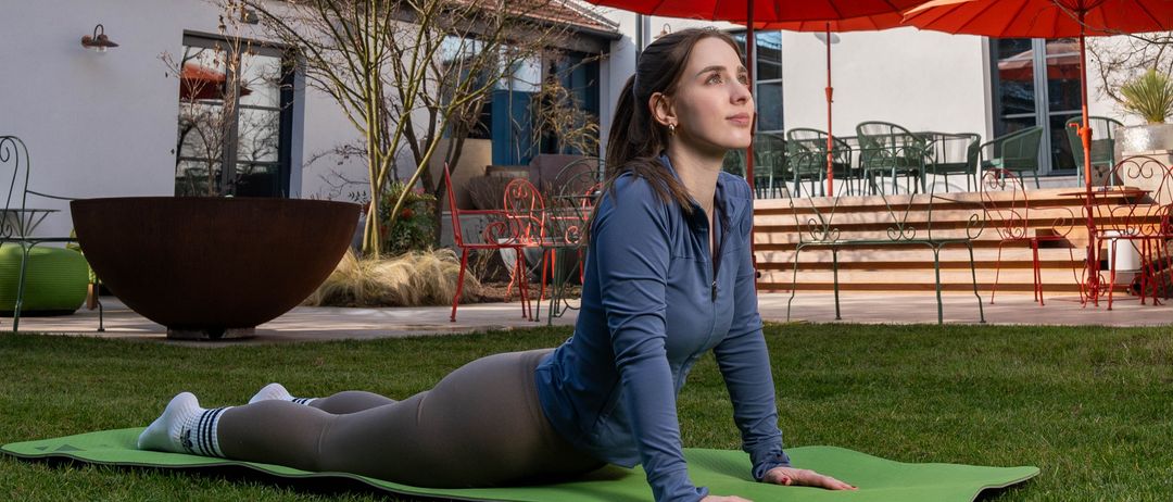 Eine Frau macht Yoga vor einem modernen Haus. Das Haus hat Glasfenster und einen Balkon. Rote Regenschirme und Möbel sind draußen.