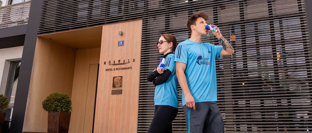 Ein Mann und eine Frau in passenden blauen Shirts stehen vor einem Gebäude mit Metallfassade und trinken aus blauen Dosen.