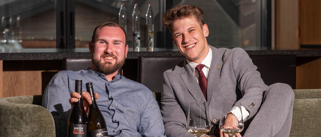 Two men sit on a couch, one in a business suit and the other in casual attire. They hold wine bottles and glasses. The man on the left wears sneakers, while the man on the right wears dress shoes. Behind them is a counter with bottles and a glass window.