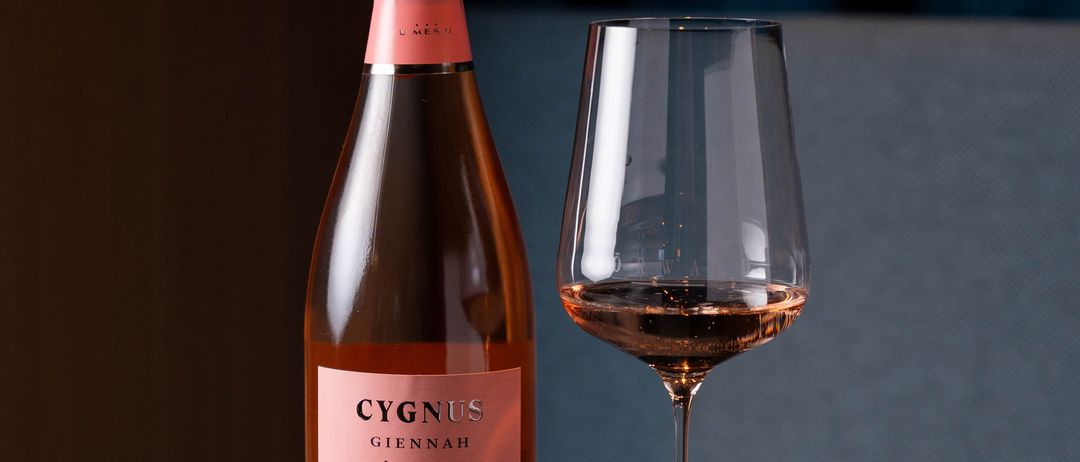 A bottle of Cygnus Giannah Brut Rose stands beside an empty wine glass on a dark wooden table.