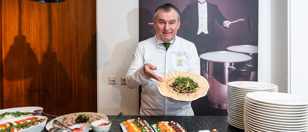 Ein Koch in einer weißen Jacke steht hinter einem Tisch mit Essen und hält einen Teller mit Gemüse. Ein großes Foto eines Mannes in einem Frack ist im Hintergrund.