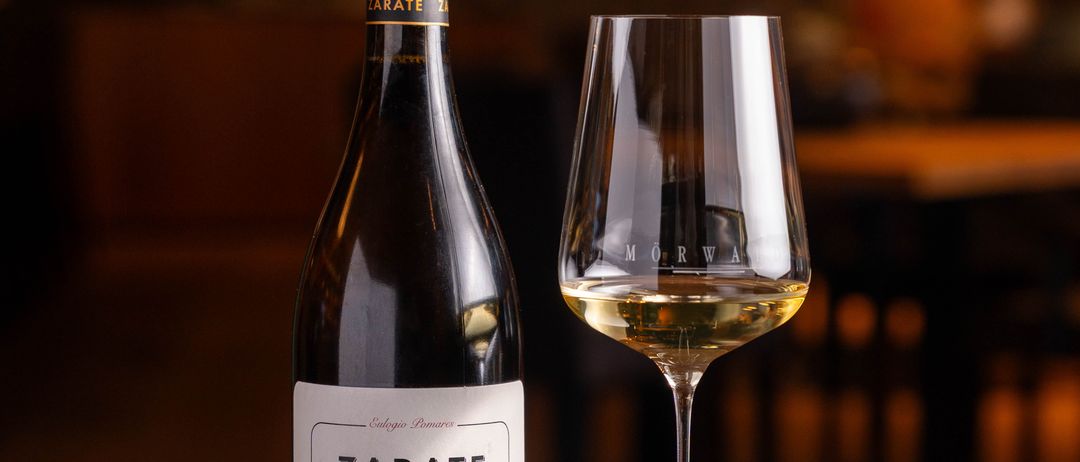 A bottle of Zarate Albariño white wine stands next to a wine glass on a wooden table, with a blurry background of a room.