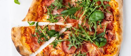 Eine ganze Pizza mit ausgeschnittenen Stücken, belegt mit Käse, Pepperoni und Rucola. Die Pizza hat einen goldbraunen Boden und steht auf einer weißen Oberfläche.