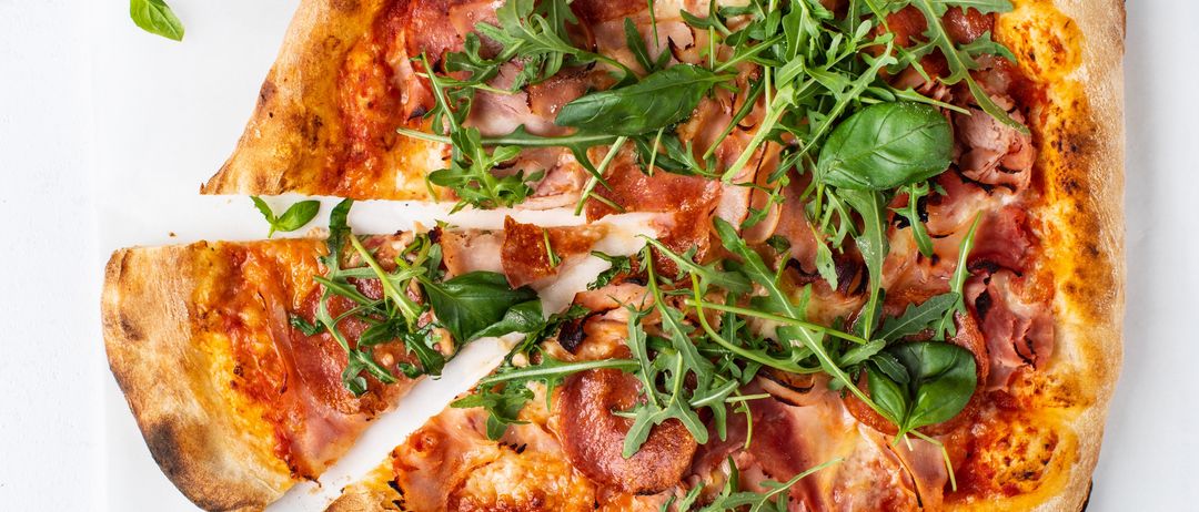 Eine ganze Pizza mit ausgeschnittenen Stücken, belegt mit Käse, Pepperoni und Rucola. Die Pizza hat einen goldbraunen Boden und steht auf einer weißen Oberfläche.