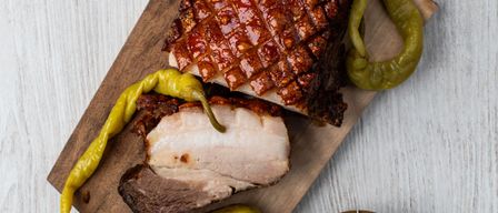 Auf einem Holzschneidebrett liegen geschnittenes, glasiertes Schweinefleisch, gelbe Paprika und ein Pickel. Ein Schüssel mit geriebenem Käse steht auf einer weißen Oberfläche.
