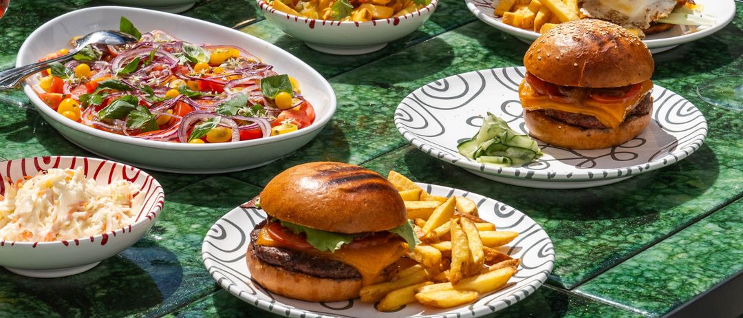 Ein Tisch mit verschiedenen Gerichten und Getränken, einschließlich Burger, Pommes, Salate und Cocktails, auf einer grünen Oberfläche mit grünen Stühlen im Hintergrund.