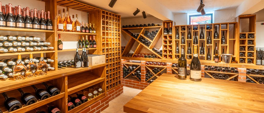 A wine cellar with wooden shelves filled with various wine bottles, illuminated by ceiling lights, and a wooden table with wine bottles on it.