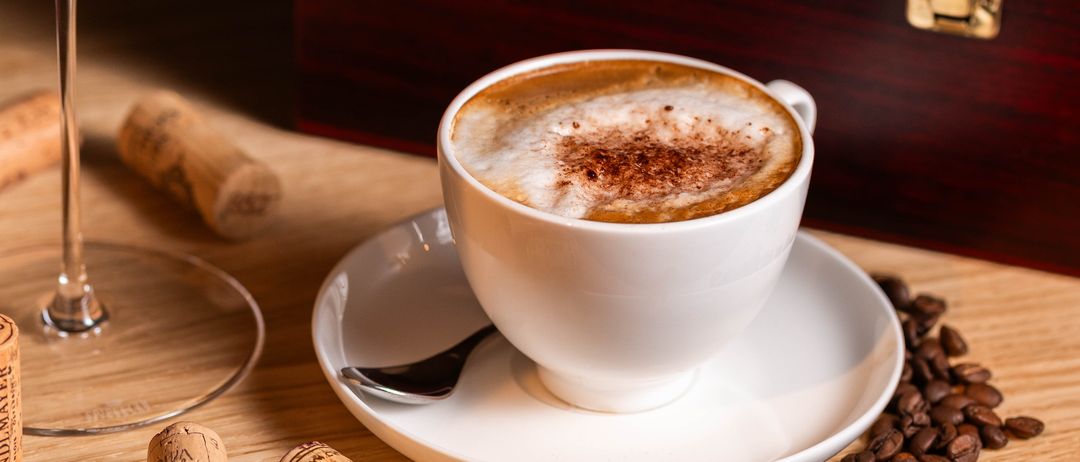 A white cup of coffee with foam and cinnamon on top, on a saucer with a spoon, on a wooden table with coffee beans and a wine cork.
