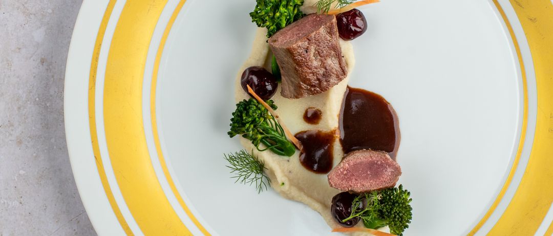 A white plate with yellow stripes holds a gourmet dish: a piece of meat with a sauce, accompanied by broccoli, a cherry, and a dollop of creamy white sauce.