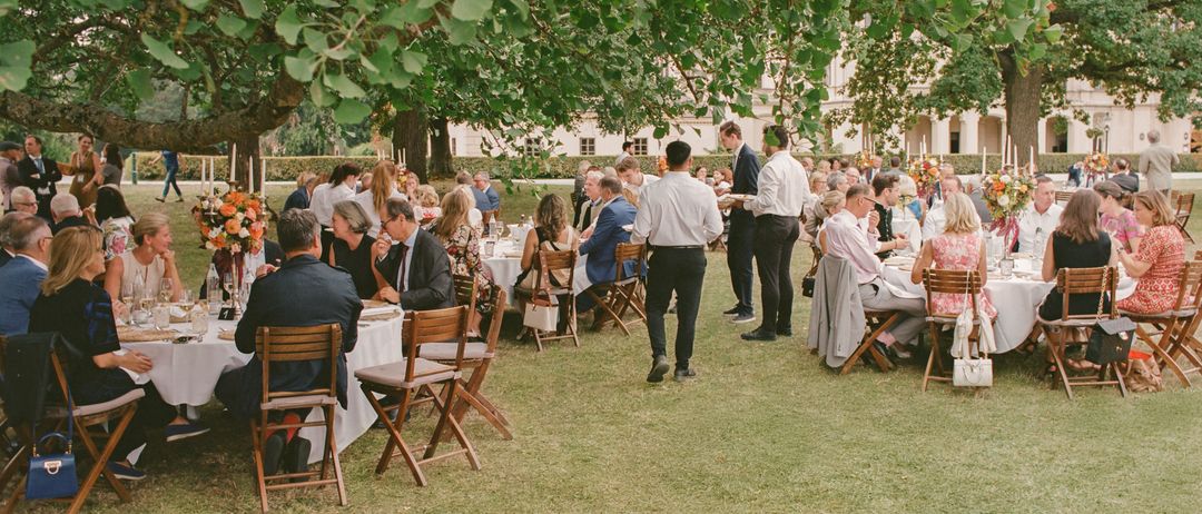 A group of people are seated at tables under trees at an outdoor event, with some standing and conversing. The tables are covered with white cloths and adorned with flowers.